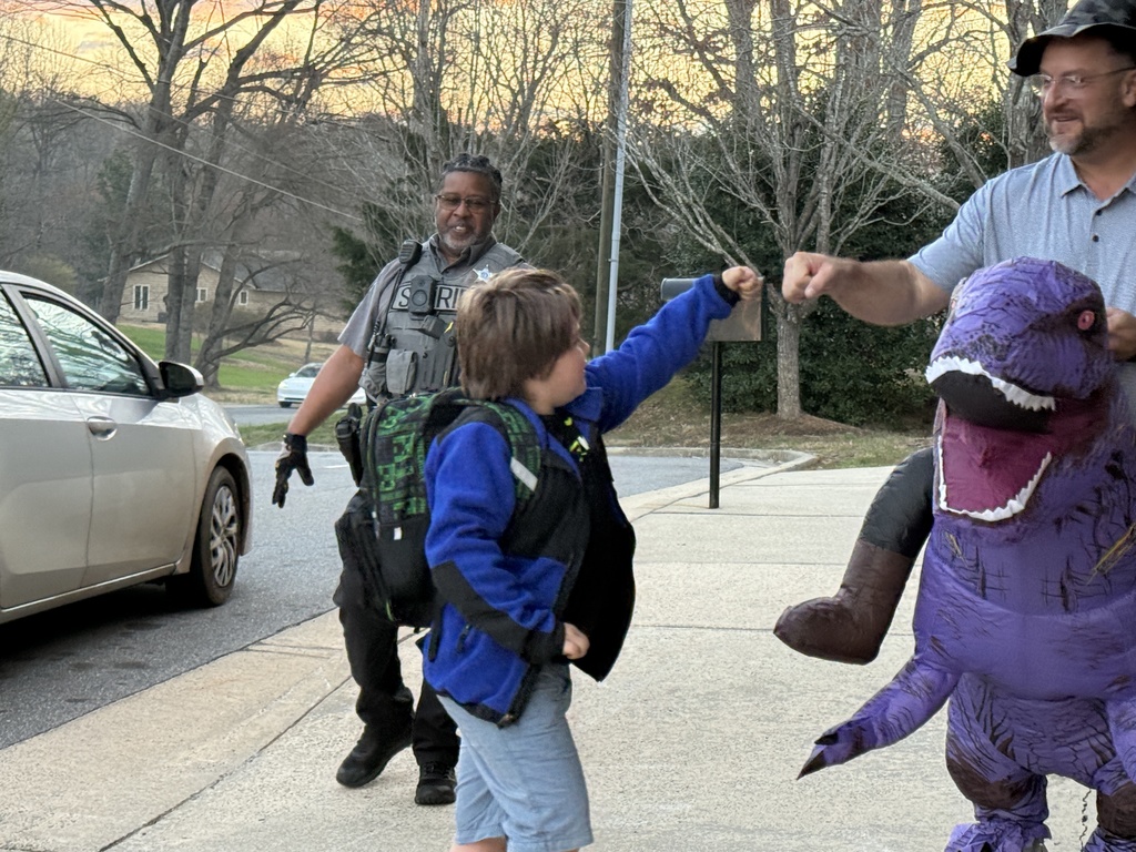 A student wearing a backpack gives a fist bump to a staff member in an inflatable purple dinosaur costume during morning arrival outside the school. A sheriff’s deputy stands nearby helping with car rider traffic while students walk toward the building.
