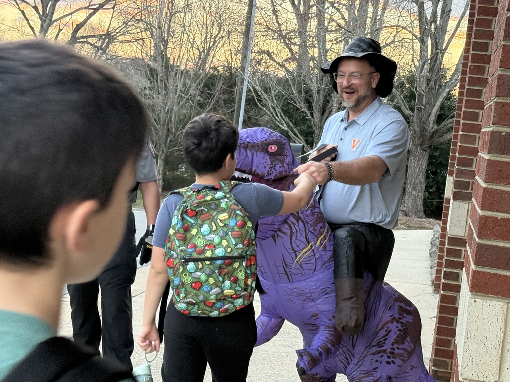A staff member wearing an inflatable purple dinosaur costume greets a student arriving at school, shaking the student’s hand as they walk toward the entrance with a backpack. The interaction takes place on the sidewalk outside the brick school building during morning arrival, with other students nearby.