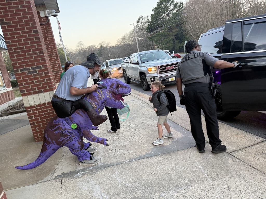 A young student laughs while interacting with a staff member in an inflatable purple dinosaur costume outside the school during morning arrival. A sheriff’s deputy assists with car rider traffic nearby while other vehicles line up to drop off students.