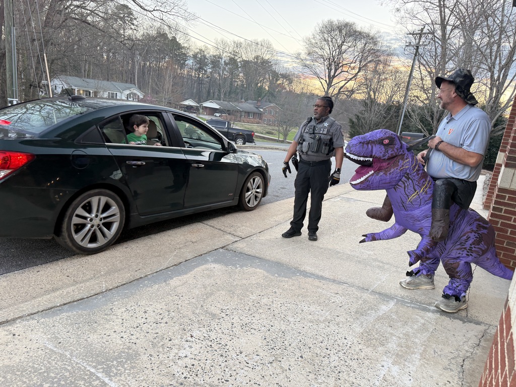 During morning car rider drop-off, a sheriff’s deputy and a staff member in an inflatable purple dinosaur costume stand on the sidewalk greeting students. A student leans out of a car window smiling while talking with them, with houses and trees visible across the street.
