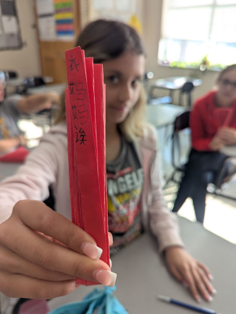 A close-up shot of a student's hand holding a folded red paper fan. Written vertically in black ink on the fan are Chinese characters. The student is blurred in the background.