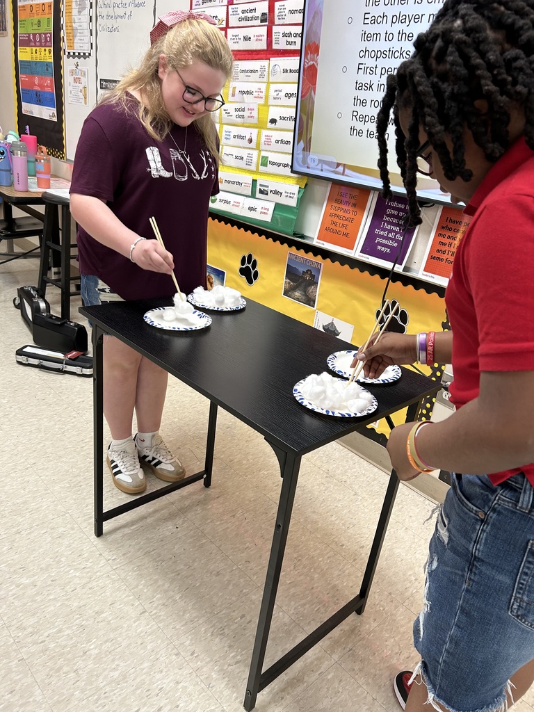 Two middle school students stand at a black table in a classroom, practicing using chopsticks to move cotton balls from one plate to another. An interactive whiteboard in the background displays information about an Ancient China unit, and classroom walls feature educational posters with vocabulary terms.