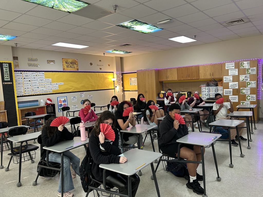 A classroom of middle school students sitting at desks, each holding a red paper folding fan in front of their face. The classroom features a yellow word wall, wooden cabinetry, and purple LED strip lighting along the walls.