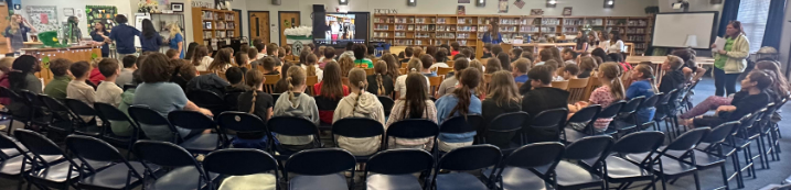 A wide-angle, back-view of a large group of students seated in rows of black folding chairs and wooden library chairs. They are gathered in a school media center or library, facing a large television screen that displays a photo of three people.