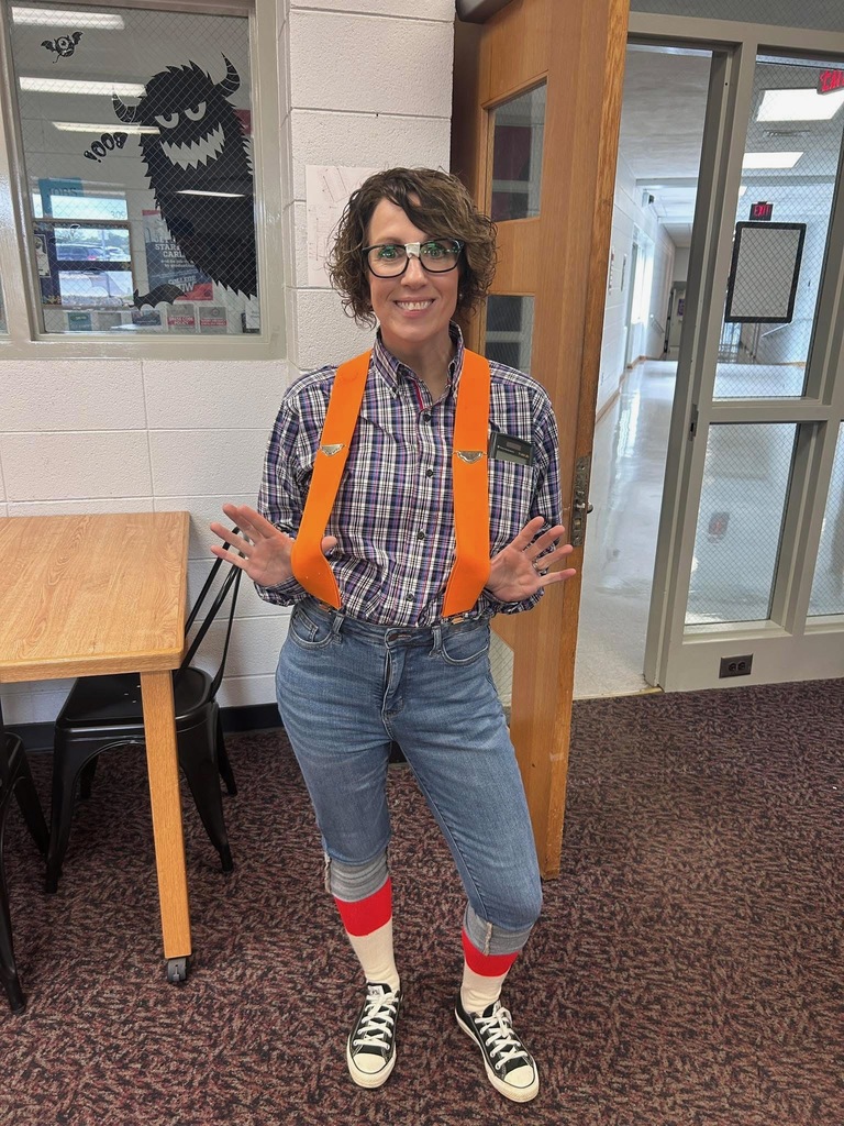 A woman with brown curly hair and glasses stands in a room with white cinderblock walls. She is dressed in a costume featuring a blue and white plaid button-down shirt, bright orange suspenders, blue jeans cuffed at the shins, and white socks with a red stripe. She is wearing black and white Converse sneakers and holding her hands up with palms facing forward. A monster decoration is visible on the window behind her.