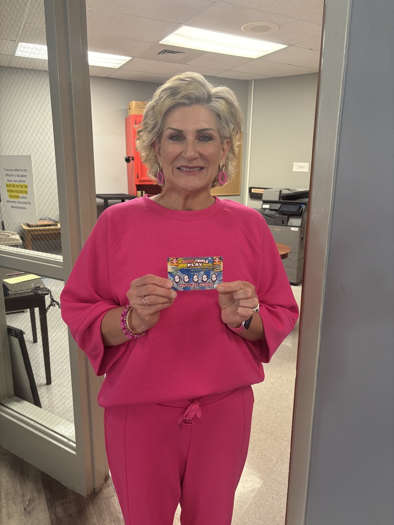A woman with blonde hair, wearing a bright pink long-sleeve sweatshirt, matching pink pants, and pink earrings, stands in a doorway. She is smiling and holding a "Triple Play" scratch-off lottery ticket with both hands. In the background, there is an office setting with a red cabinet, a printer, and a sign on the glass door regarding cell phone usage in the office.
