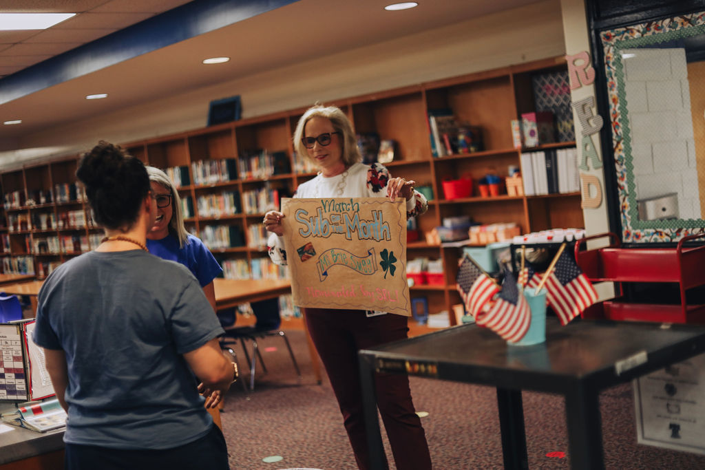 A woman presents a handmade poster reading “March Sub of the Month – Ms. Brie Sugg – Nominated by SRL!” to a staff member in a school media center. Two other women stand nearby as they share the recognition moment. Bookshelves and a table with small American flags are visible in the foreground.