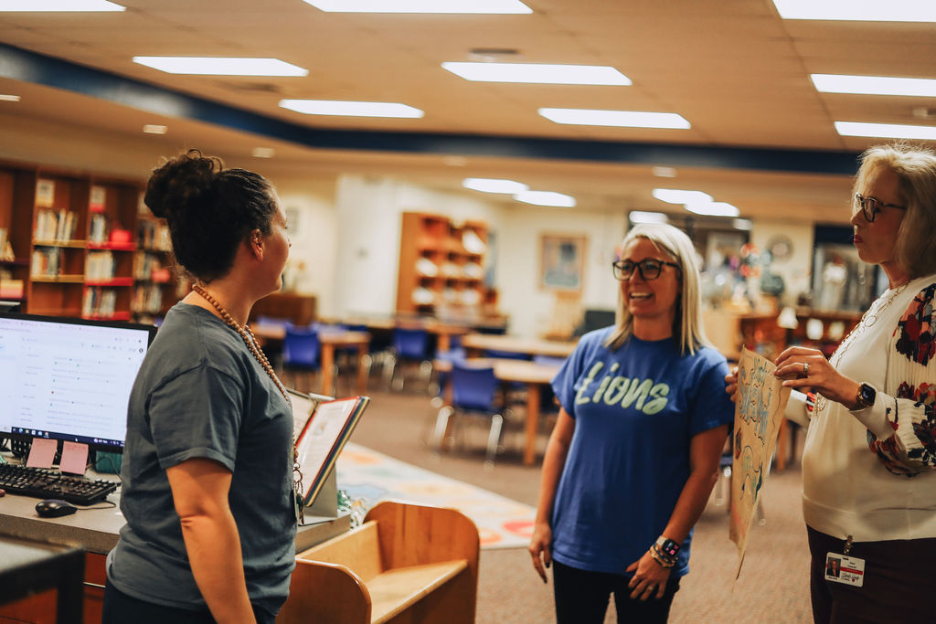 Three women stand near the circulation desk of a school media center during a conversation. One woman faces the other two while they smile and talk, holding a handmade poster recognizing “March Sub of the Month – Ms. Brie Sugg – Nominated by SRL!”