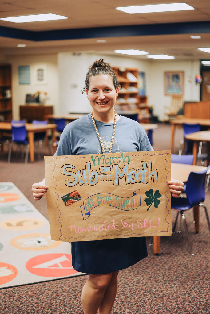 A smiling woman stands in a school media center holding a handmade poster that reads “March Sub of the Month – Ms. Brie Sugg – Nominated by SRL!” The poster is decorated with colorful lettering, a small image of pizza, and a green shamrock. Bookshelves, student tables, and blue chairs fill the background of the library.