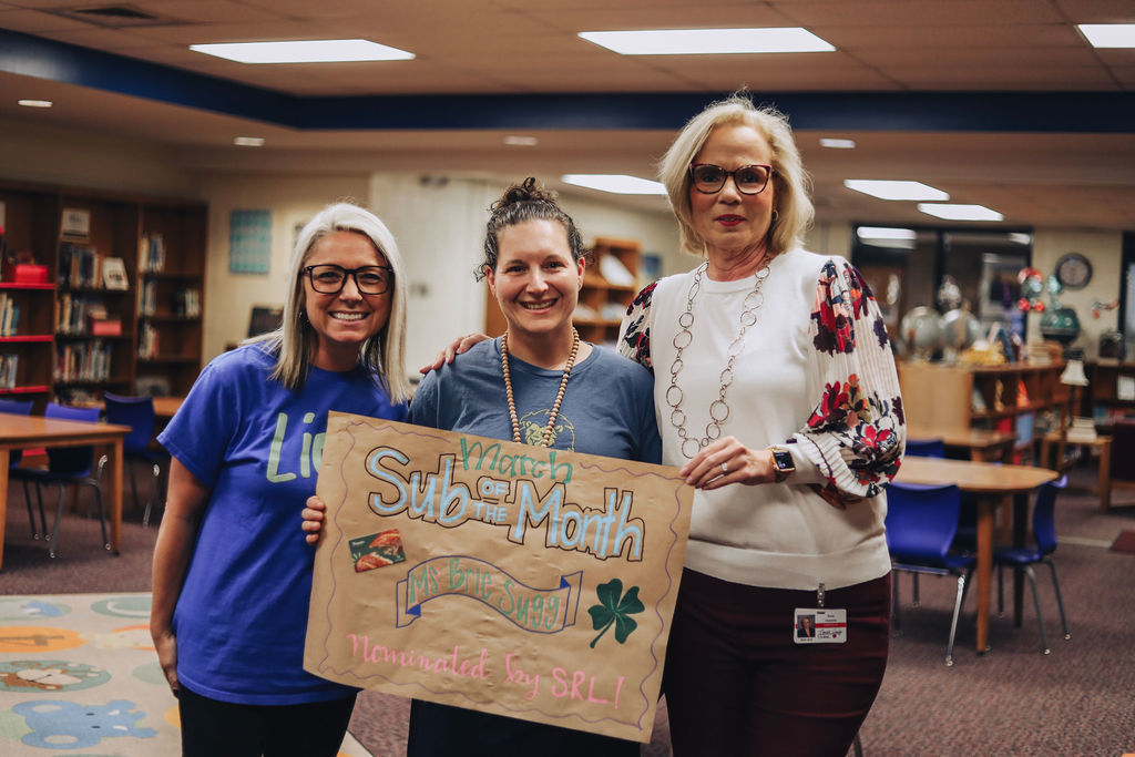 Three women stand together in a school media center holding a handmade poster that reads “March Sub of the Month – Ms. Brie Sugg – Nominated by SRL!” The woman in the center holds the sign while the two women beside her smile for the photo. Bookshelves and student tables are visible behind them.
