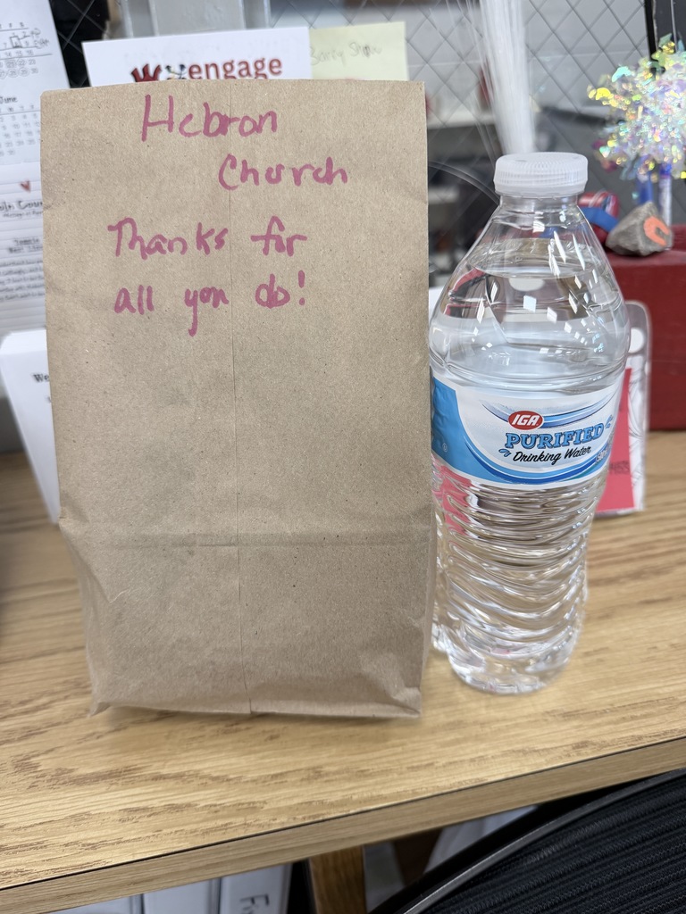 A brown paper lunch bag sits on a wooden desk next to a clear plastic bottle of IGA Purified Drinking Water. Handwritten in pink marker on the bag is the text: Hebron Church Thanks for all you do!