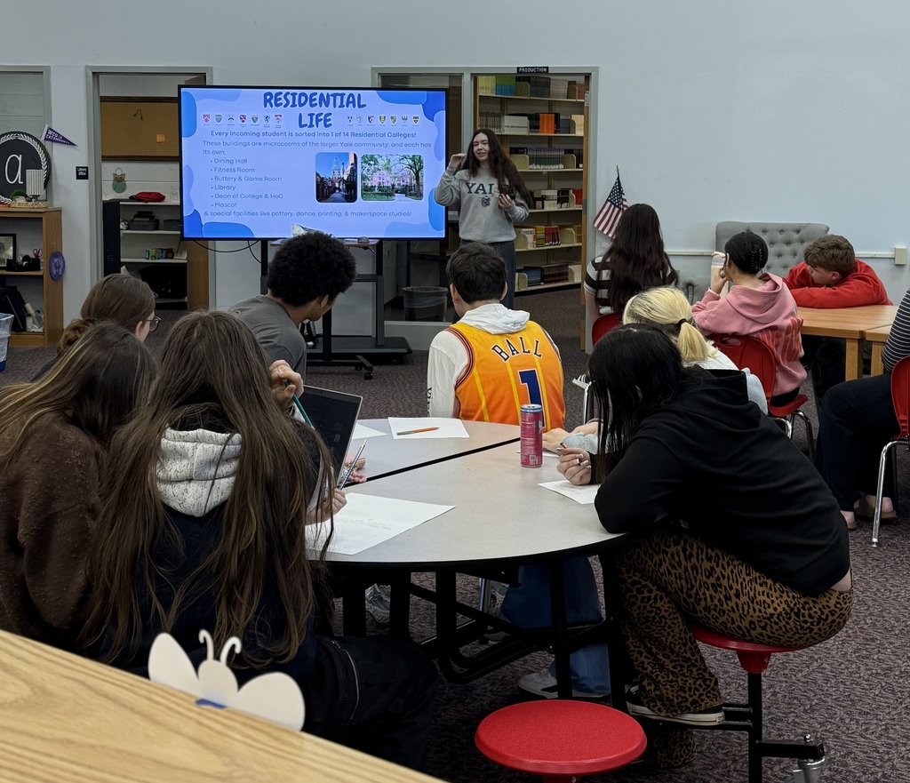 A group of high school students sit at a round table in a media center, listening to a presentation about Yale University's residential life. A presenter in a Yale sweatshirt stands in the background by a large screen. One student in the foreground wears an orange basketball jersey with the name BALL and the number 1 on the back.