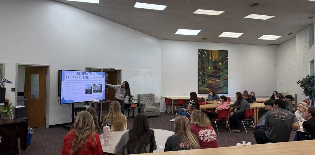 A wide shot of a school media center where a presenter is pointing to a digital screen titled RESIDENTIAL LIFE. Approximately twenty students are seated at various tables, listening to the presentation. A large piece of art depicting a whimsical forest path hangs on the back wall.