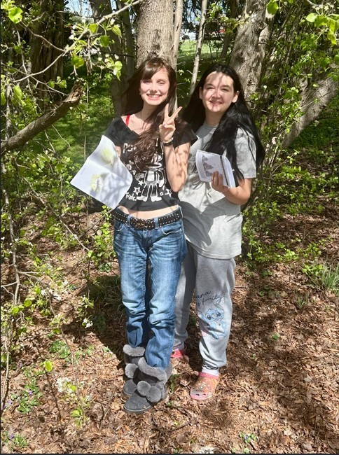 Two female students stand together outdoors among trees with small white blossoms. The student on the left wears a black t-shirt, blue jeans, and grey fur-lined boots, holding a paper and making a peace sign. The student on the right wears a grey t-shirt and grey sweatpants featuring a cartoon character, also holding a paper. They are standing on a bed of dry leaves and mulch.