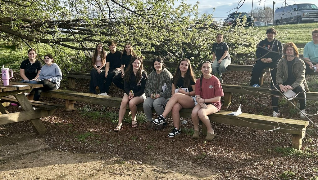 A group of approximately fifteen students are gathered outdoors in a shaded seating area made of wooden benches. They are sitting under the low-hanging branches of a large tree filled with white blossoms. Many students are holding small white booklets or papers, and a picnic table is visible on the far left.