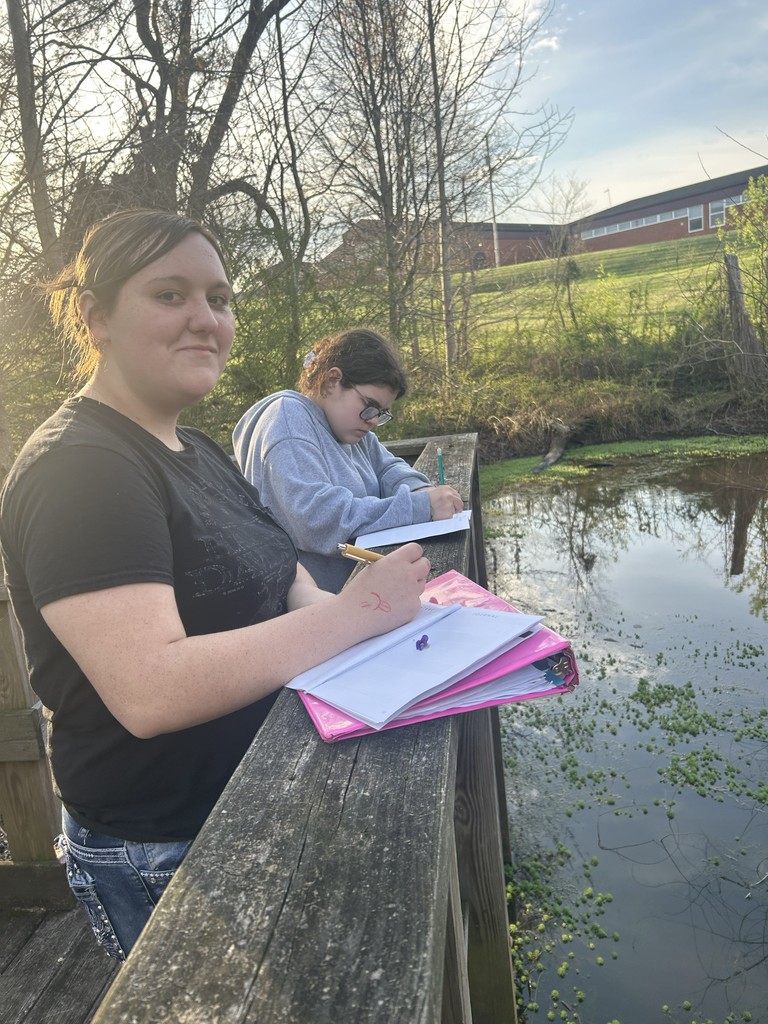 Two students stand on a wooden boardwalk overlooking a pond. The student in the foreground wears a black t-shirt and has a pink binder open on the railing, while the student in the background wears a grey hoodie and glasses while writing on a notepad. The pond contains green aquatic plants, and a school building is visible on a hill in the distance.