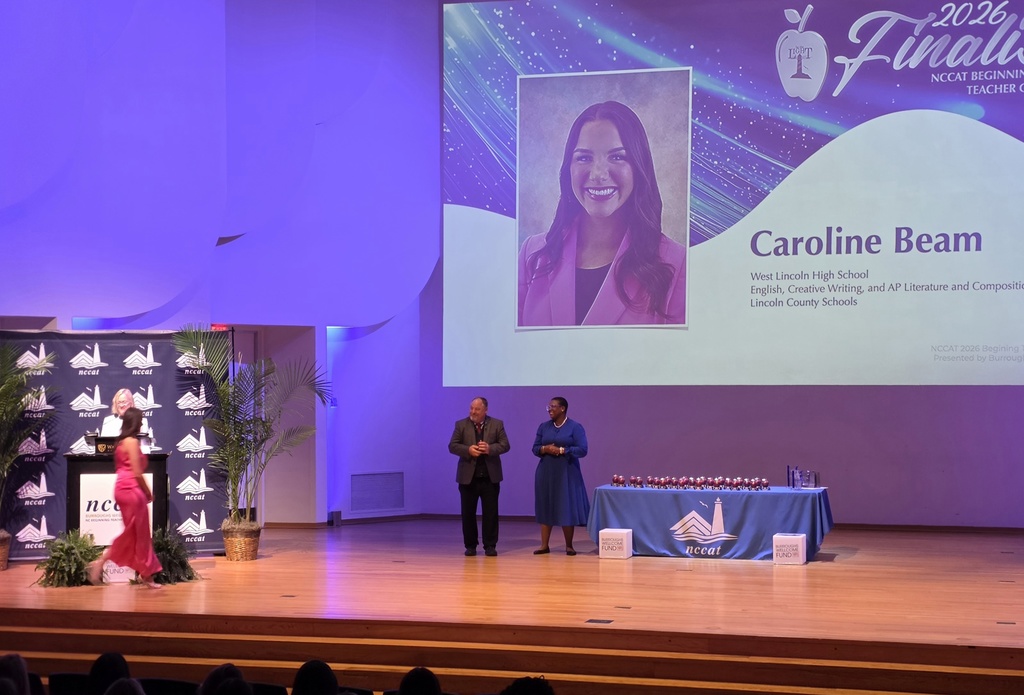 A large stage at the NCCAT ceremony displays a presentation slide with a portrait of Caroline Beam and text identifying her as a teacher at West Lincoln High School teaching English, Creative Writing, and AP Literature and Composition. Tables with apple-shaped awards sit on the stage while presenters stand nearby.