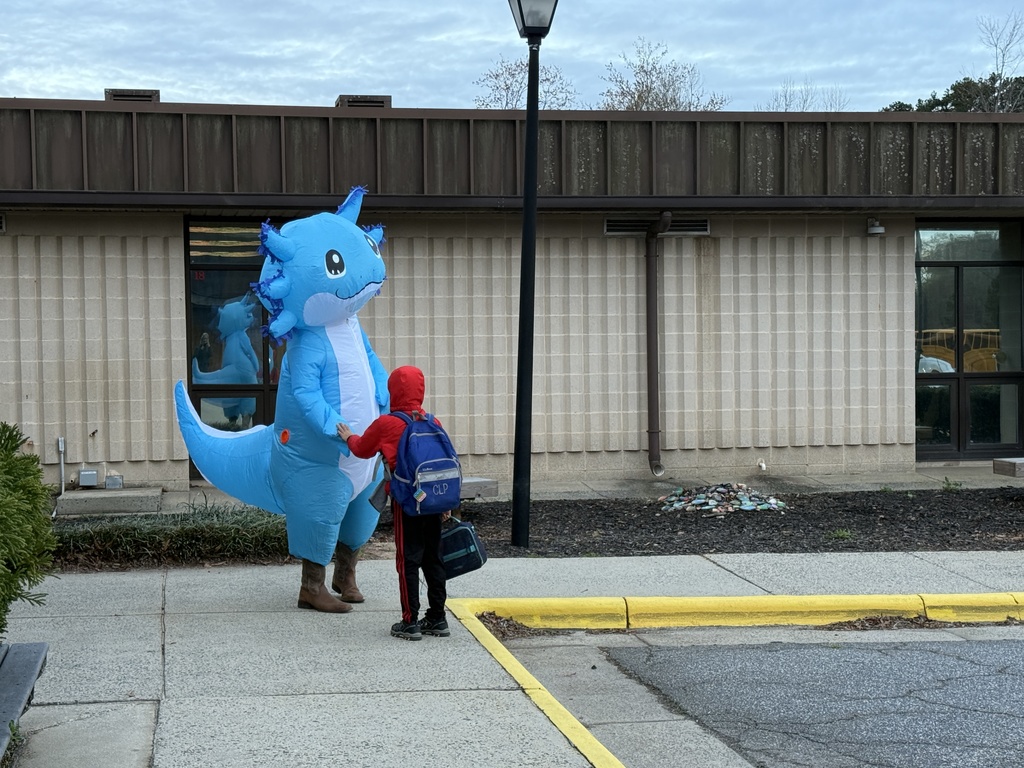 A student with a backpack stands on a sidewalk outside a school building, interacting with a person in a bright blue inflatable axolotl costume during morning drop-off.