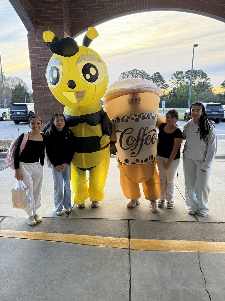 Four elementary-aged students stand outside a school entrance smiling beside two people in inflatable costumes—a large yellow-and-black bee and a giant coffee cup—during morning arrival.