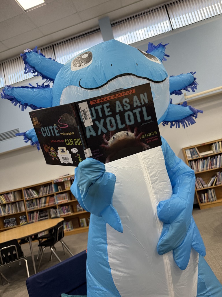 A person in a blue inflatable axolotl costume stands in a school library holding open a book titled “Cute as an Axolotl,” with bookshelves and tables visible in the background.
