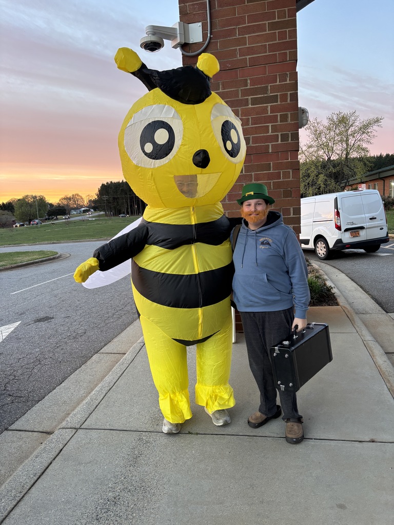 A student wearing a green hat and orange beard stands next to a person in a large inflatable bee costume outside a school at sunrise, holding a small briefcase.