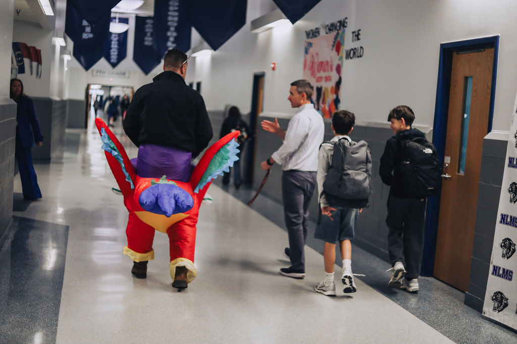 Dr. Allen, wearing an inflatable parrot rider costume, walks down a middle school hallway with two students while a staff member talks with them.