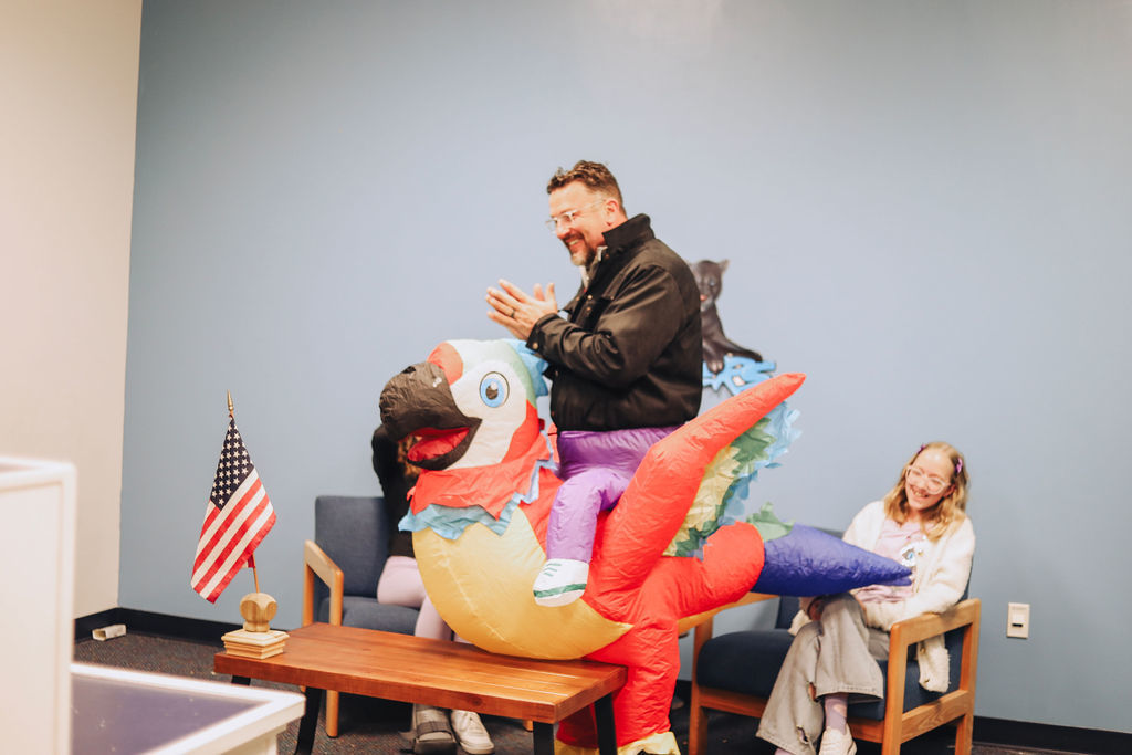 Dr. Allen, wearing an inflatable parrot rider costume, smiles and claps while seated in a school office area. A student sitting nearby laughs as she watches.