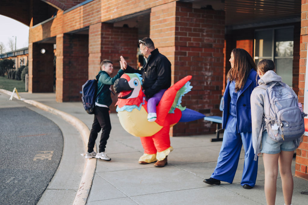 Dr. Allen, wearing an inflatable parrot rider costume, gives a student a high five outside during morning arrival as two other students stand nearby.