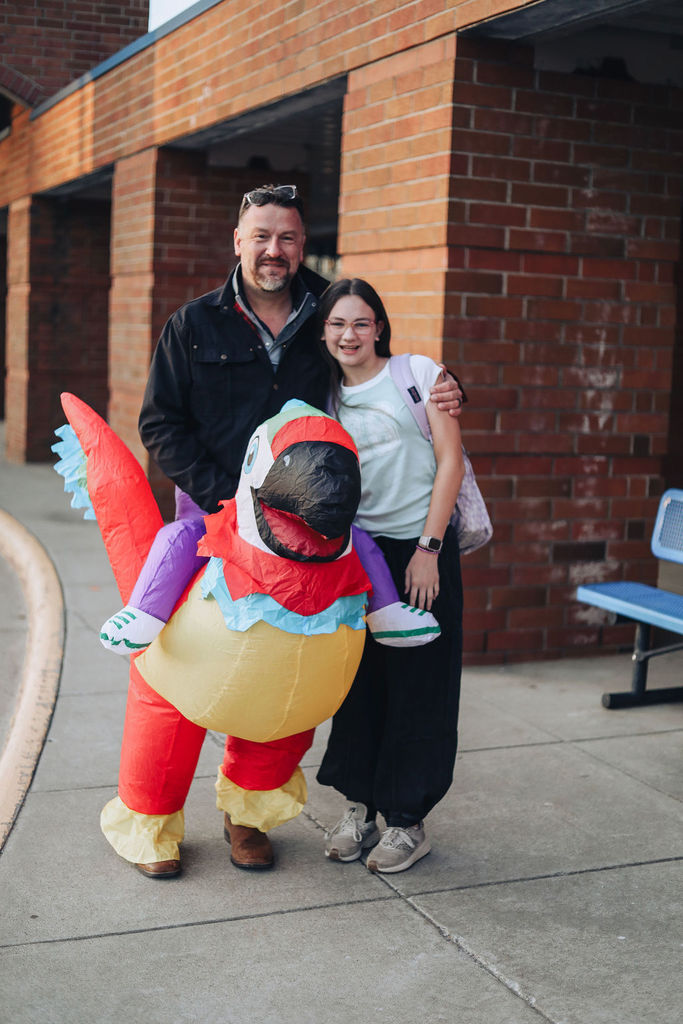 Dr. Allen stands outside a school entrance with a smiling student while wearing an inflatable parrot rider costume.