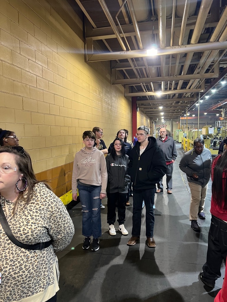 This photo shows the group continuing their walking tour through a long corridor of the industrial facility. Detailed Image Description Foreground: On the far left, the woman in the leopard-print hoodie is partially visible in profile. The shadows of the photographer and other group members are cast on the dark grey concrete floor in the lower center. Middle Ground: The students and chaperones are walking in a loose line toward the back of the facility. Visible members include a young woman in a tan "Aero" hoodie and ripped jeans, a young woman in a black "Champion" hoodie, and the young man with the patterned buzz cut wearing a black Carhartt jacket. All continue to wear clear safety glasses. Further back, an older man in a grey pullover and several other individuals follow behind. Background: To the left is a tall, yellow cinder block wall. Above, a complex system of industrial pipes, conduits, and light fixtures runs the length of the ceiling. To the right, various pieces of machinery and yellow safety railings line the walkway, extending deep into the background of the factory floor. Setting: The perspective emphasizes the scale of the facility, with the long lines of the wall and ceiling pipes creating a sense of depth. The lighting is bright and industrial.