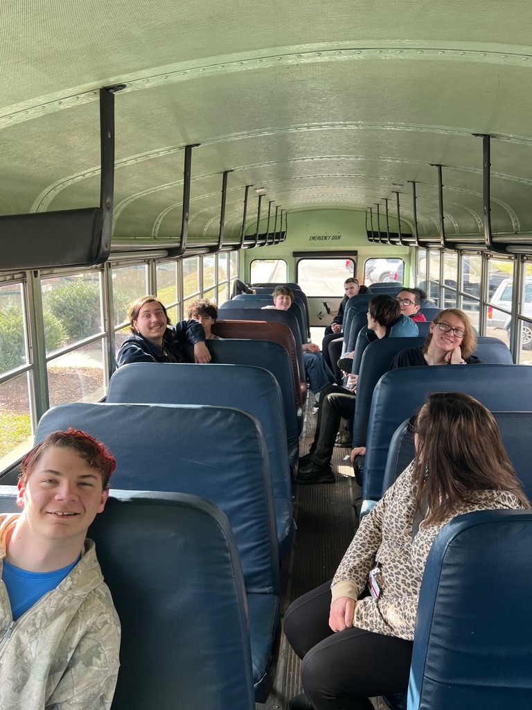 This photo captures a group of people sitting on a school bus. The perspective is from the front of the bus looking towards the back, showing the interior with its characteristic rows of blue padded seats and pale green walls and ceiling. Detailed Image Description Foreground: On the left, a young man with light skin and short brown hair with red highlights smiles at the camera. He wears a camouflage hoodie over a blue shirt. On the right, a person with long, dark wavy hair is seen from the back/side, wearing a leopard-print zip-up hoodie. Middle Ground: Several rows of seats are filled with young adults. On the left side, a young man with dark hair and a mustache smiles broadly, leaning against the seat. Further back, others are scattered throughout the bus, some looking toward the camera and others looking away. Background: The aisle leads to the back of the bus, where an "EMERGENCY DOOR" sign is visible. Through the windows on both sides, a parking lot and some green bushes can be seen under bright, natural daylight. Setting: The bus interior features metal luggage racks running along both sides of the ceiling and large rectangular windows that fill the space with light.