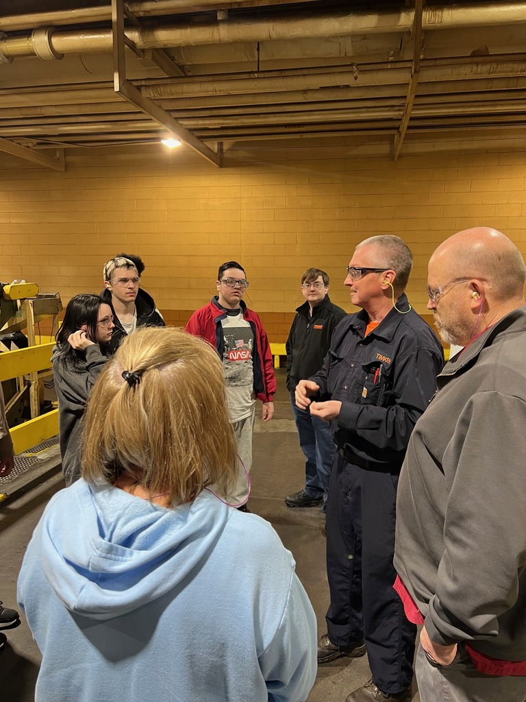 This photo shows a group of young adults gathered in a circle inside an industrial plant, listening to an employee. Detailed Image Description Foreground: In the lower center, a person with blonde hair tied in a small ponytail wears a light blue hoodie, seen from the back. To the right, an older man with a grey beard and glasses listens intently, also seen partially from the side. Middle Ground: An employee, a middle-aged man with short grey hair and glasses, stands in the center-right. He wears a navy blue TIMKEN work shirt, an orange undershirt, and yellow earplugs. He is gesturing with his hands as he speaks. The Group: Surrounding the speaker are several young adults from the previous photos. They are all wearing clear safety glasses and earplugs. Notable individuals include the young man in the red jacket and NASA t-shirt and the young man with the patterned buzz cut. Background: The setting is the factory floor, featuring a yellow cinder block wall and a high ceiling with exposed pipes and industrial light fixtures. To the left, a piece of heavy machinery with bright yellow safety railings is visible. Setting: The lighting is bright and functional. The group's focused expressions suggest a high level of engagement during the tour.