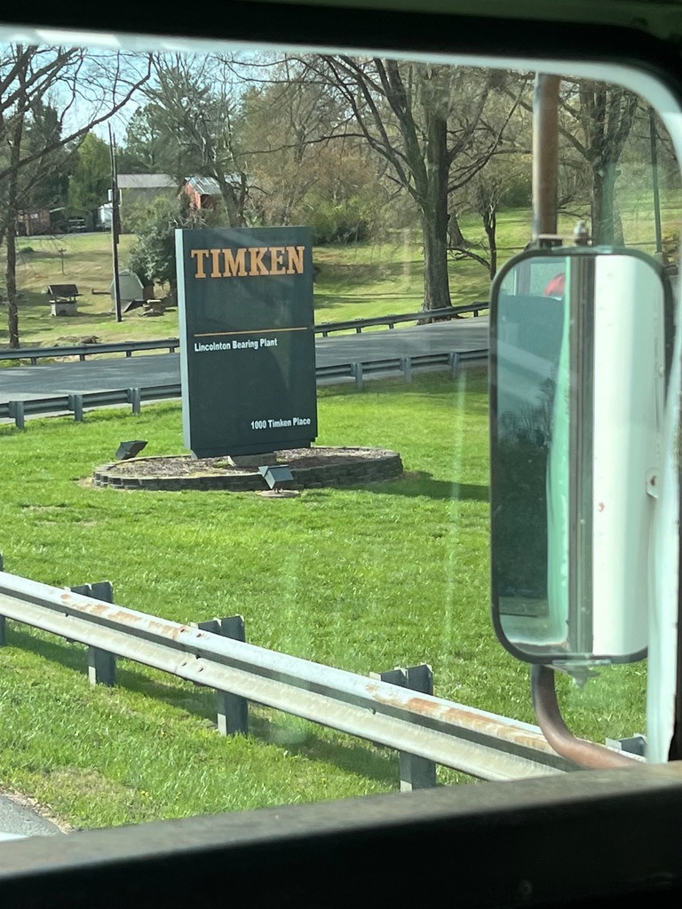 This image is a view from inside a vehicle, likely a bus, looking out of the passenger-side window at an industrial sign. Detailed Image Description Foreground: The bottom and right edges of the frame show the interior window seal and a large, vertical side-view mirror of the vehicle. The Sign: In the center of the frame stands a tall, dark green rectangular sign for TIMKEN. The company name is at the top in large, orange-gold block letters. Below a thin horizontal line, smaller white text reads "Lincolnton Bearing Plant" and "1000 Timken Place" at the very bottom. The sign sits in a small circular mulch bed bordered by grey stones on a green lawn. Background: Beyond the sign is a paved road lined with a metal guardrail. In the distance, there are rolling green hills, a small wooden structure (possibly a well or shed), and several leafless deciduous trees, suggesting early spring or late fall. Setting: The scene is brightly lit by daylight, capturing a suburban industrial area in Lincolnton.