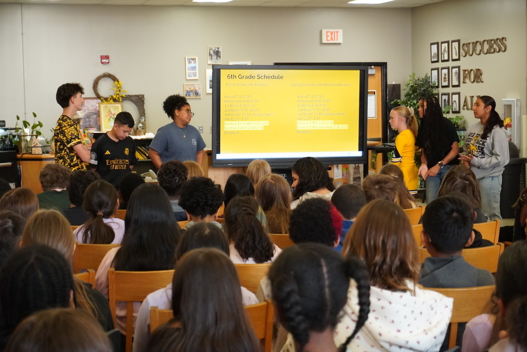 6th Grade Schedule. Fall Schedule with Electives: Arrival 7:15-7:50, Homeroom 8:00-8:30, 1st Block 8:30-9:35, 2nd Block 9:35-10:40, 3rd Block 10:40-12:15 (include lunch), 4th Block 12:15-1:15, Elective 1 (Health) 1:25-2:10, Elective 2 (Art) 2:10-2:55, Dismissal. Spring schedule with Different Electives: Arrival 7:15-7:50, Homeroom 8:00-8:30, 1st Block 8:30-9:35, 2nd Block 9:35-10:40, 3rd Block 10:40-12:15 (include lunch), 4th Block 12:15-1:15, Elective 1 (PE) 1:25-2:10, Elective 2 (Keypad/Grit) 2:10-2:55, Dismissal. Five students stand around the large monitor displaying this schedule in a media center or classroom setting.