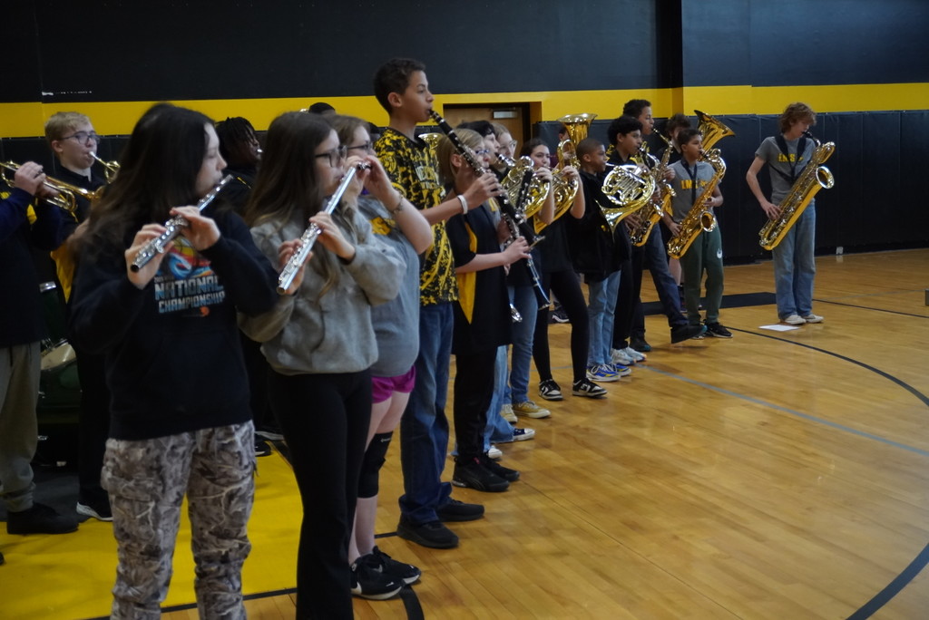 A group of middle school band students stands in a gymnasium, playing various instruments. In the foreground, two girls play silver flutes. Behind them, a boy plays a clarinet, and further back, students play brass instruments including French horns and a baritone saxophone. The students are dressed in a mix of casual clothes and school-colored yellow and black apparel.