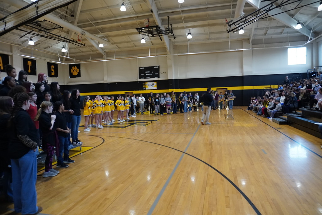 A wide-angle view of a school gymnasium during an assembly. On the left, a choir stands on tiered risers, and a line of cheerleaders in yellow and black uniforms stands on the court. In the center, a band is lined up, and a staff member in a black shirt and khaki pants walks across the hardwood floor. On the right, a large audience of students and adults sits in the bleachers.