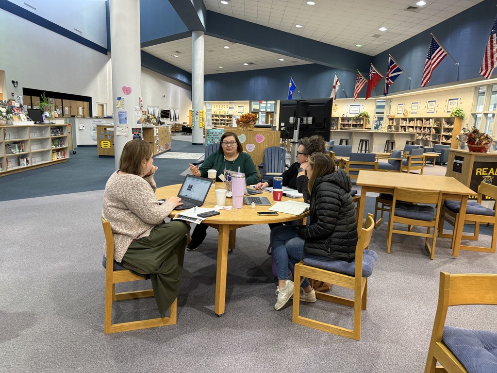 Four staff members sit around a round table in a spacious school media center, talking and working on laptops. Coffee cups, notebooks, and papers are spread across the table, and the open library space with bookshelves, seating areas, and flags is visible in the background.