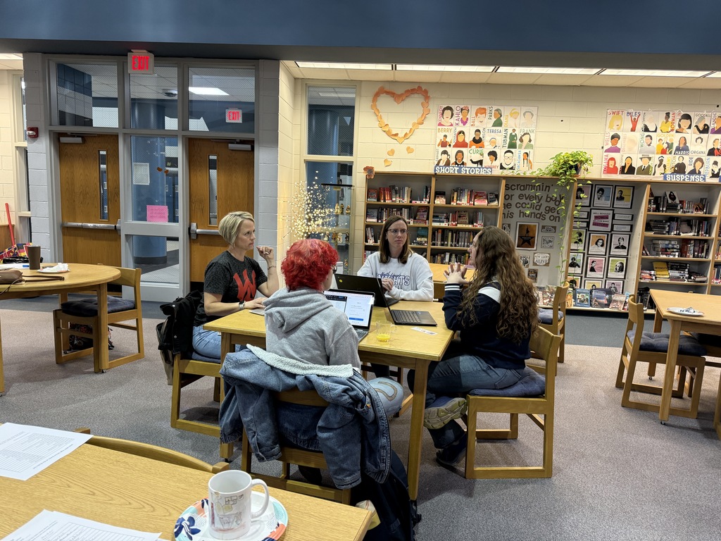 Four educators sit around a table in a school library, engaged in discussion with laptops open in front of them. Behind them are bookshelves, student artwork, posters featuring authors and genres, and a display labeled “Short Stories.”