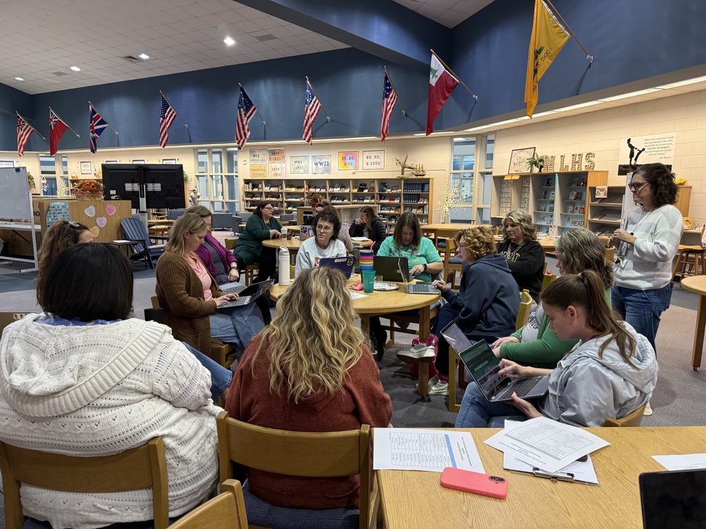 A large group of educators sits in a circle and at round tables in a high school media center during a collaborative meeting. Several staff members use laptops while one woman stands and speaks to the group. Bookshelves, school decor, and multiple flags line the walls of the spacious library.