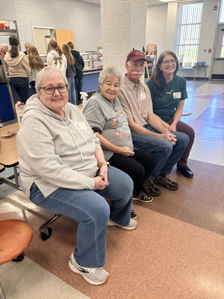 Four adults sit together on a bench in a school cafeteria setting. On the far left, a woman with short gray hair and glasses wears a gray hooded sweatshirt and blue jeans. Next to her, an older woman with short dark hair wears a gray t-shirt with a floral graphic and the words "God all things are Possible." Beside her is a man with a mustache wearing a maroon baseball cap and a striped button-down shirt. On the far right, a woman with long brown hair and glasses wears a green t-shirt with an "nrg" logo and a name tag. In the background, students and staff are visible near a cafeteria serving line and large windows