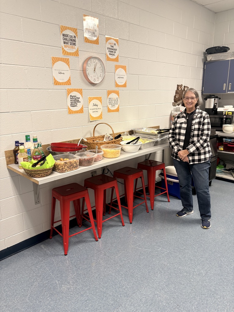 A woman with short gray hair and glasses, wearing a black and white buffalo plaid shirt and blue jeans, stands next to a salad bar setup in a room with white cinderblock walls. The salad bar is arranged on a counter with four red metal stools tucked underneath. It includes various toppings in clear containers, salad dressings, a basket of croutons, and large silver catering pans. On the wall above the counter, a circular clock is surrounded by several orange and white motivational flyers with text including "Magic lies in challenging what seems impossible," "Unity is the strongest form of magic," and "Always believe that something wonderful is about to happen.