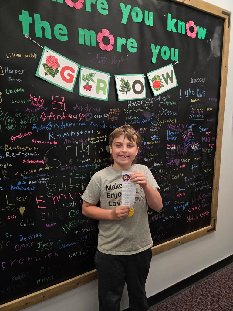 A young boy in a grey t-shirt stands in front of a black signature wall, proudly holding up a long, narrow slip of paper containing a list of completed math multiplication problems. The wall behind him features a teal "the more you know the more you GROW" display and is covered in names written in colorful chalk.