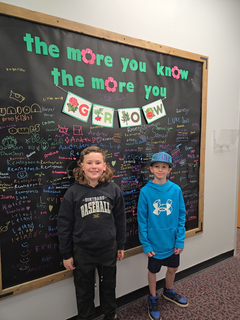 Two young boys stand side-by-side in front of a large black signature wall framed in wood. The boy on the left wears a black "Dirtbags Baseball" hoodie, and the boy on the right wears a bright blue Under Armour hoodie and a blue baseball cap. Above them, a teal title reads "the more you know the more you GROW." The wall is densely covered with names handwritten in multi-colored chalk.