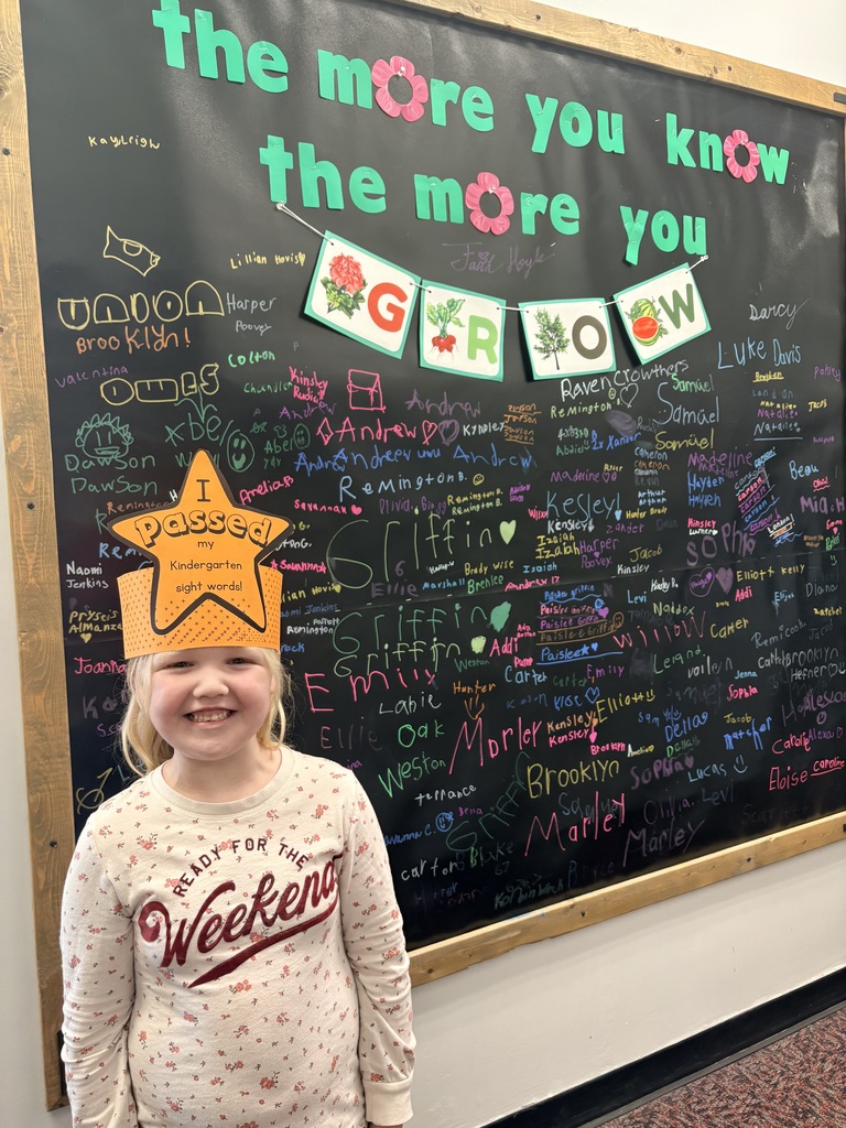 A young girl with blonde hair and a floral long-sleeved shirt smiles while wearing an orange paper achievement crown that reads "I Passed my Kindergarten sight words!" She is positioned in front of a "the more you know the more you GROW" bulletin board. The black background behind her is filled with dozens of student names written in various bright chalk colors.