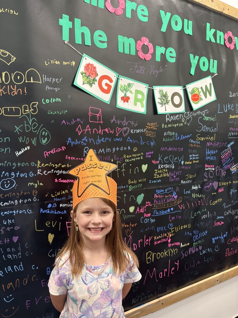 A young girl with long reddish-brown hair smiles broadly while wearing an orange paper crown that says "I Passed my Kindergarten sight words!" She is standing in front of a large black chalkboard wall covered in colorful handwritten names. Above her, a teal sign reads "the more you know the more you GROW" with a small banner showing illustrations of plants.