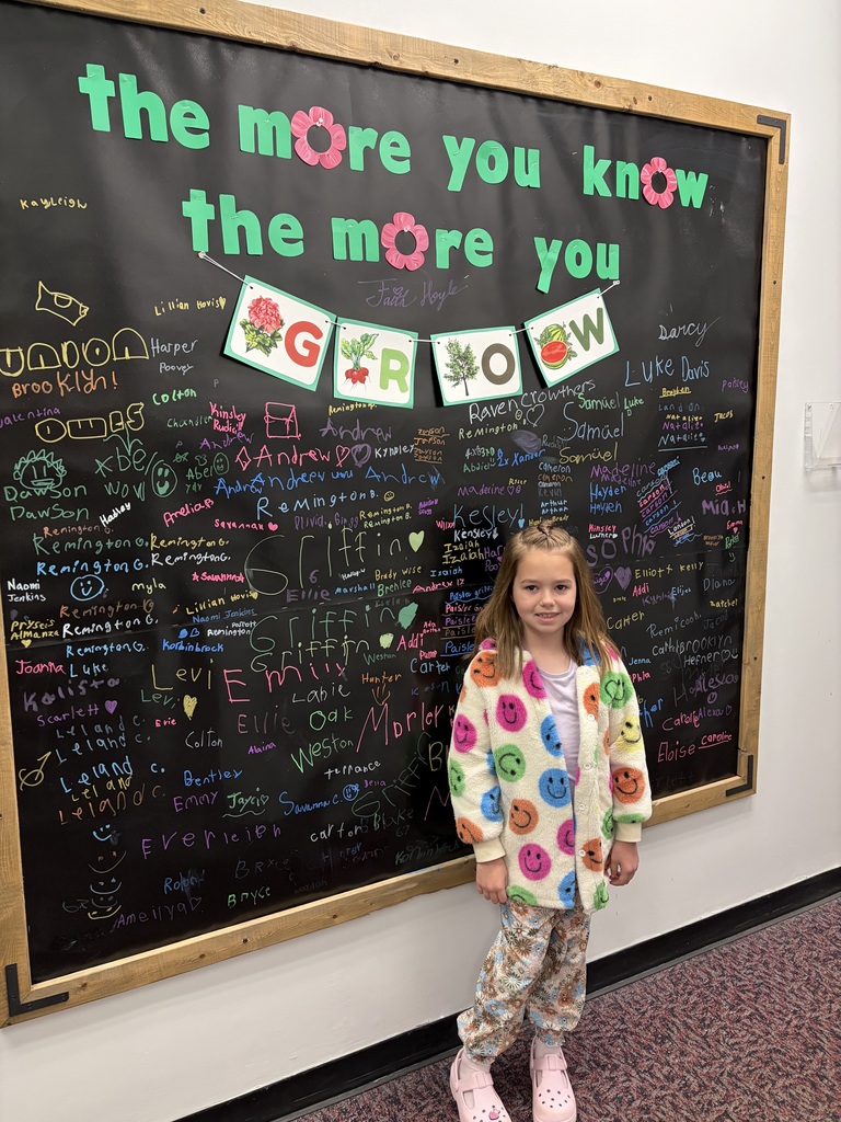 A young girl with light brown hair stands in front of a large black chalkboard signature wall. She is wearing a cream-colored fuzzy jacket covered in multi-colored smiley faces, floral print pants, and pink clogs.  Above her, a teal sign reads "the more you know the more you GROW," decorated with pink paper flowers and a small banner illustrating plants. The chalkboard background is filled with dozens of student names handwritten in various bright chalk colors.