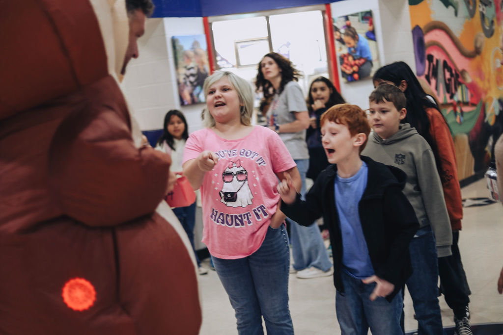 Students stand in a school hallway interacting with a person in a large inflatable costume. Two elementary-aged students in the foreground point and react with excitement, while other students and an adult watch in the background near colorful wall murals and classroom displays.