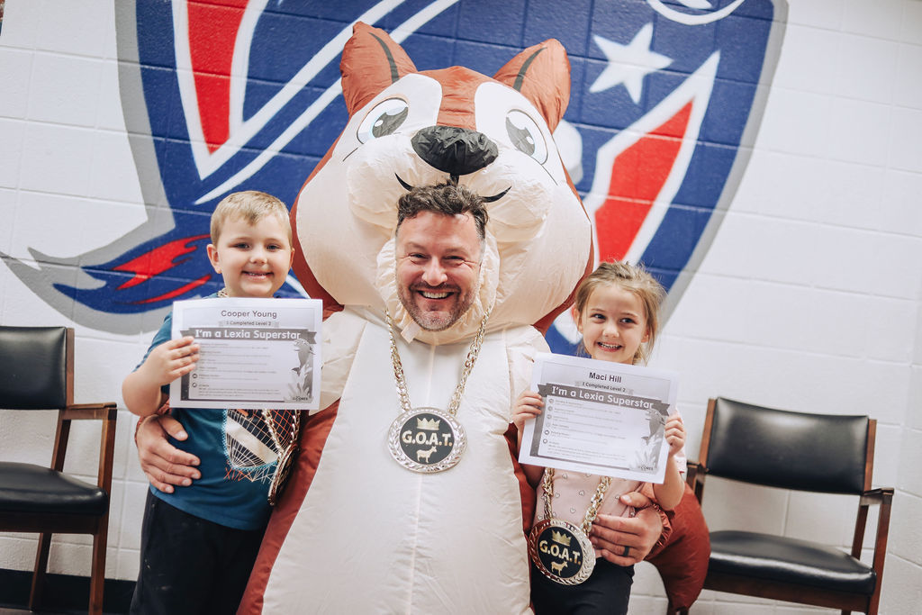 An adult wearing an inflatable animal costume and a large “G.O.A.T.” medallion kneels between two young students holding certificates that read “I’m a Lexia Superstar.” All three are smiling, seated in front of a school wall with a large painted mascot logo.