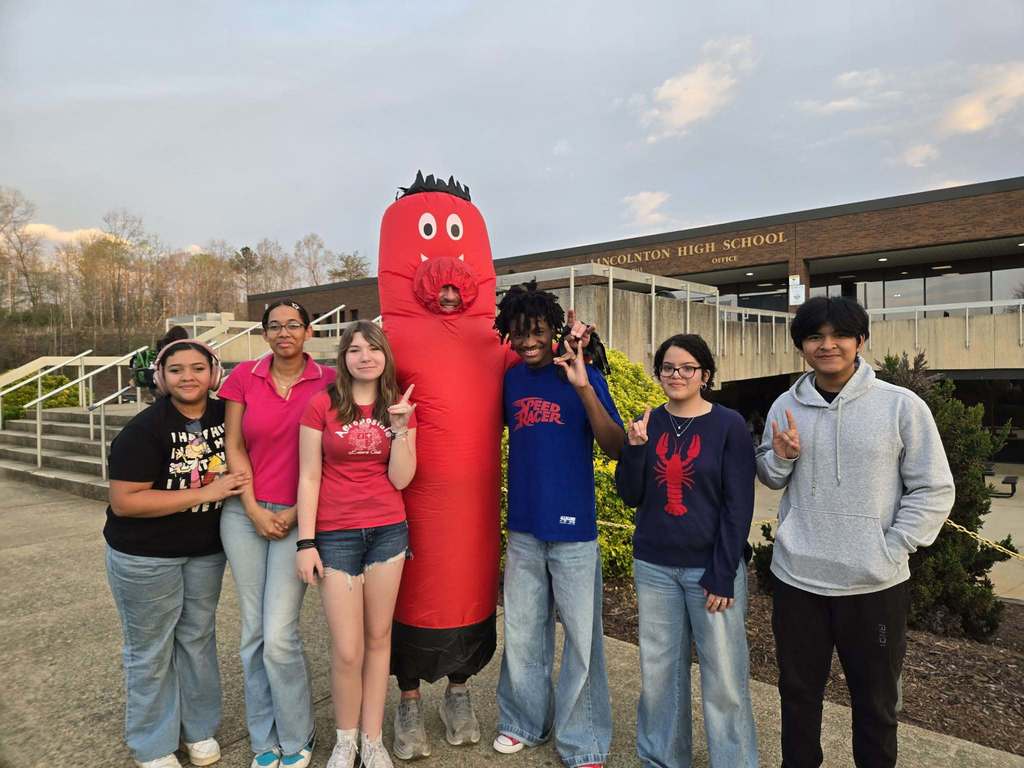 A group of high school students stands outside Lincolnton High School posing with a person in a tall red inflatable costume. The students smile and gesture with their hands, standing on a sidewalk near the school entrance with the building and landscaping visible behind them.