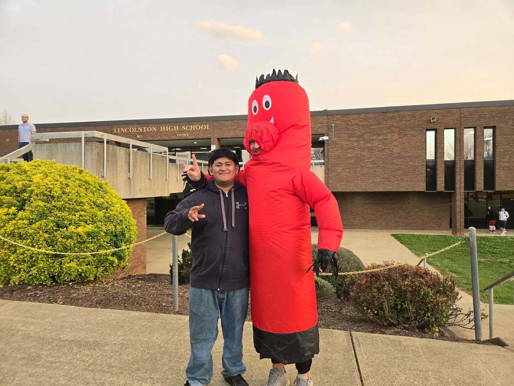 A high school student stands next to a person in a tall red inflatable costume outside Lincolnton High School. Both pose with hand gestures in front of the school building, with shrubs, walkways, and the school sign visible in the background.